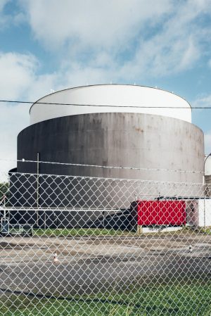Ein großer, zylindrischer Industrietank hinter einem Maschendrahtzaun. Ein roter Container steht im Vordergrund.
