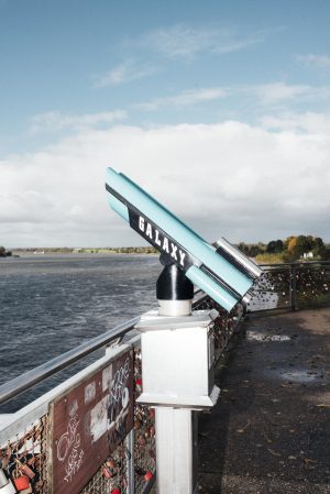 Ein türkisfarbenes Fernglas mit der Aufschrift „Galaxy“ auf einer Brücke in Wesel, mit Blick auf den Rhein, umgeben von Liebesschlössern.