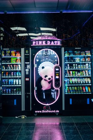 Ein Spielautomat mit der Aufschrift „Pink Date“ in Neonlichtern, umgeben von Snack- und Getränkeautomaten. Im Inneren des Automaten befindet sich ein überdimensionales Plüschpanda-Kostüm.