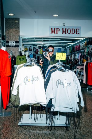Ein Mann mit Kamera steht in einem Bekleidungsgeschäft mit SALE-Schild. Vor ihm hängen Pullover mit der Aufschrift „Perfect“.