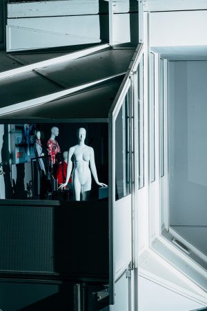 Eine Schaufensterpuppe in einem Schaufenster mit kalter Beleuchtung. Im Hintergrund sind weitere Puppen und eine Spiegelung zu sehen.