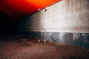 A dark underpass with a red-lit ceiling and a stained concrete wall. A graffiti tag reading "Banksy" is visible on the wall.