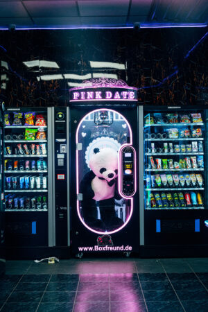 A vending machine labeled "Pink Date" in neon lights, surrounded by snack and drink dispensers. Inside the machine is an oversized plush panda costume.