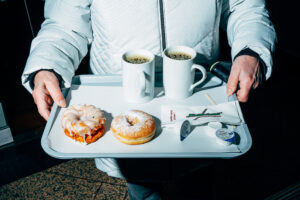 A tray with two cups of coffee and pastries, held by a person wearing a white winter jacket. The pastries include a powdered sugar donut and a glazed fried ring.