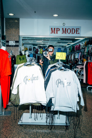 A man with a camera stands inside a clothing store with a SALE sign. In front of him, sweatshirts with the word "Perfect" are displayed.