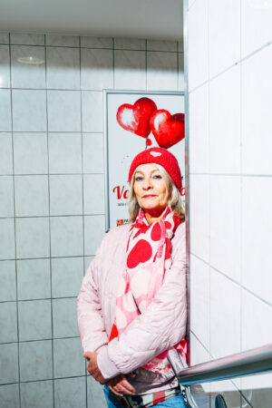 An older woman in a pink outfit with heart motifs leans against a white tiled wall. Behind her, a poster with red heart balloons is visible.