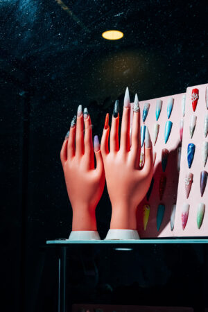 Two artificial hands with long, decorated nails stand in a store window. Next to them, a color palette displays various nail designs.