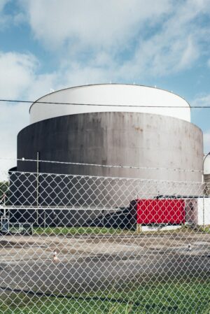 A large cylindrical industrial tank behind a chain-link fence. A red container stands in the foreground.