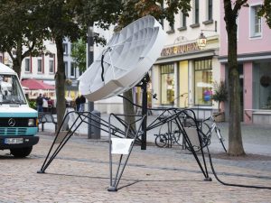 Eine futuristische Satellitenschüssel auf spinnenartigen Beinen in einer Innenstadt.