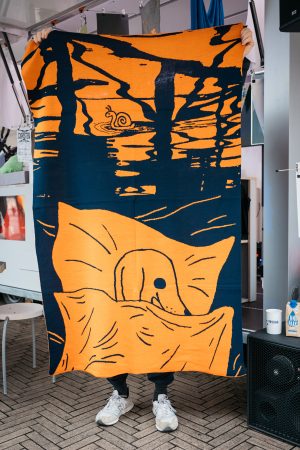 A person holds up a large orange-blue blanket with a drawing of a snail and a dog on it.
