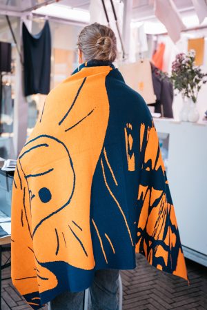 Eine Person mit zusammengebundenen Haaren trägt eine orange-blaue Decke mit Schneckenmotiv über den Schultern.