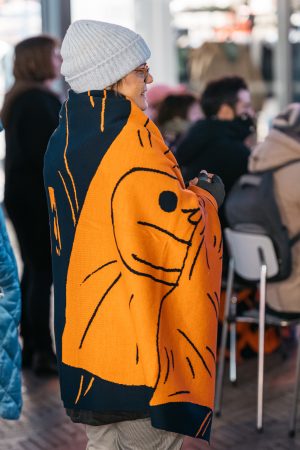 Eine Person mit Mütze und Brille steht in einer Gruppe und trägt eine orange-blaue Decke mit Schneckenmotiv um sich.