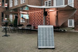 Nahaufnahme einer mobilen Bühne mit einer Leinwand und Lautsprechern. Im Vordergrund ein Aufsteller mit einer geschwärzten Zeitung.