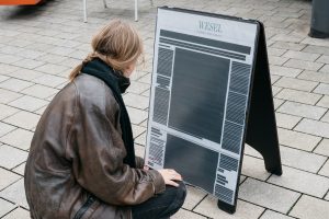Eine Person mit brauner Lederjacke betrachtet eine große, gedruckte Zeitung auf einem Aufsteller mit der Überschrift 'Wesel'.