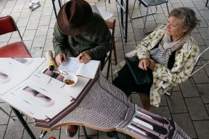 Ein älterer Mann mit Hut schreibt in ein Notizbuch an einem Tisch mit gedrucktem Architekturmotiv.