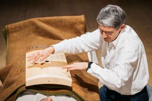 A person in a white shirt plays a string instrument over another person covered with a blanket.