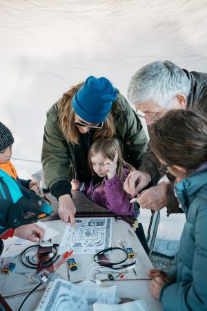 Children and adults work together on an electronics DIY project under a white cover.
