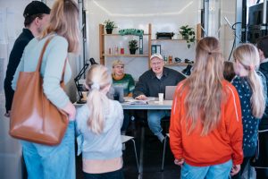 Autor Tim Holland und Autorin Caren Jeß sitzen im Trailer der Grand Snail Tour an einem Tisch und sprechen mit einer Gruppe von Kindern und Erwachsenen, die vor dem Bürotisch stehen.
