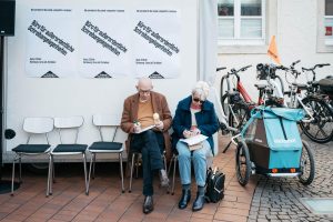 Zwei ältere Menschen sitzen vor dem Trailer der Grand Snail Tour und füllen Aufträge für ihren Wunschtext aus. Neben ihnen stehen Fahrräder.