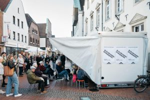 Der Trailer der Grand Snail Tour dient als mobiles Büro bei der Veranstaltung in Haltern am See. Eine Gruppe von Menschen versammelt sich auf der Fußgängerzone, einige sitzen unter der ausgeklappten Plane des Trailers.