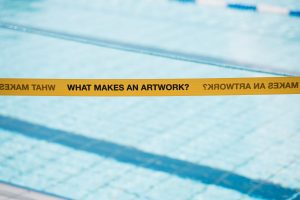 Gelbes Absperrband von Cem A. mit der Aufschrift „WHAT MAKES AN ARTWORK?“ vor dem Wasser des Solebads.