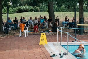 Publikum sitzt im Schatten der Bäume am Solebad und hört der Veranstaltung zu; im Vordergrund das gelbes Hinweisschild von Cem A.