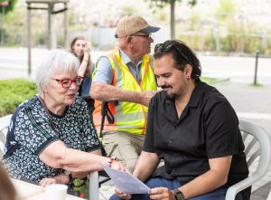 Driant Zeneli sitzt mit einer Zeitzeugin an einem Tisch und spricht mit ihr, im Hintergrund weitere Besucher*innen der Grand Snail Tour.