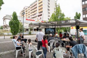 Besucher*innen sitzen vor dem mobilen Trailer und verfolgen die Filmvorführung und Gespräche über das ehemalige City-Hochhaus.