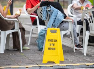 Zwischen den sitzenden Besucher*innen steht ein gelbes Warnschild mit der Aufschrift „DO NOT TALK TO THE ARTWORK“, ein Teil des Kunstwerks von Cem A.