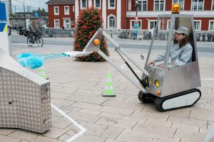 A person operates a small artistic mini-excavator with a blue duster attachment, part of the SONDER Hammer Caretaker Championships course.
