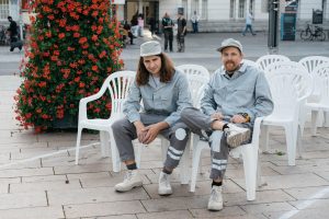 The two artists of SONDER sit in grey work uniforms on white chairs within the Hammer Caretaker Championships course.
