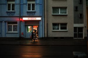 Photograph by Fatih Kurçeren: Two people with an umbrella walk past a brightly lit kiosk in the evening.