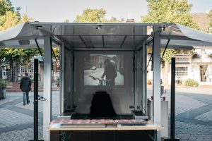 A photo by Fatih Kurçeren is projected inside the open trailer; in front of it is a small stage with a table and technical equipment.