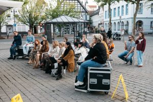 Audience members sit on chairs and equipment cases in the square, watching the event in front of the trailer.
