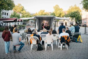 The audience sits in front of the open trailer; Esra Canpalat reads on the stage, with a projection by Fatih Kurçeren in the background.