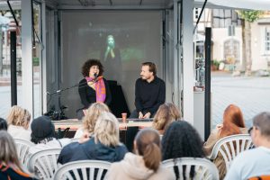 Esra Canpalat and Julian Rauter speak on the trailer stage; behind them is a projected photo by Fatih Kurçeren.