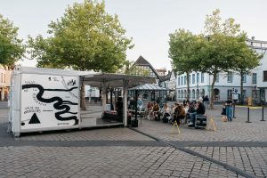 The trailer with its open stage stands on the square; in front of it the audience listens to Esra Canpalat’s reading.