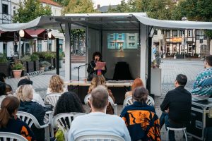 Audience members sit in front of the trailer watching the reading; a projected photograph by Fatih Kurçeren appears in the background.