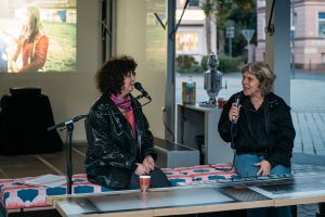 Esra Canpalat sits on the stage talking with Britta Peters; behind them a projection by Fatih Kurçeren is visible.
