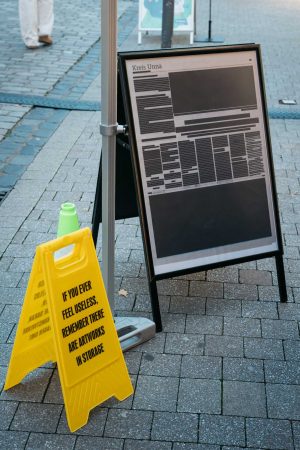 Die geschwärzte Zeitung „Local Blackout“ von Lütfiye Güzel steht in einem Aufsteller neben dem gelben Warnaufsteller aus Cem A.s „floor pieces and stanchions“.