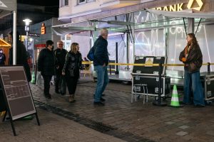 Passant*innen gehen an der Station der Grand Snail Tour vorbei; im Trailer ist die Laserinstallation „spin jump crawl climb dream bite hunt“ von Paul Spengemann zu sehen, eingerahmt vom Absperrband von Cem A. und einem Aufsteller von Lütfiye Güzel.