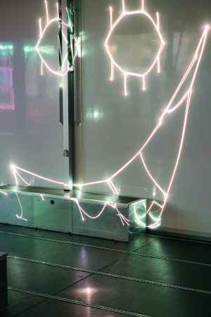 Nahaufnahme der Laserlinien von Paul Spengemanns „spin jump crawl climb dream bite hunt“, die sich über die weiße Wand und eine Metallbox im Trailer ziehen.